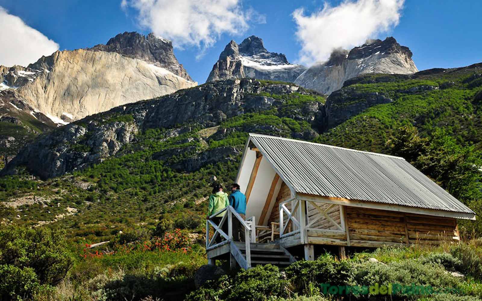 Two people standing in front of a cabin with mountains in the background.