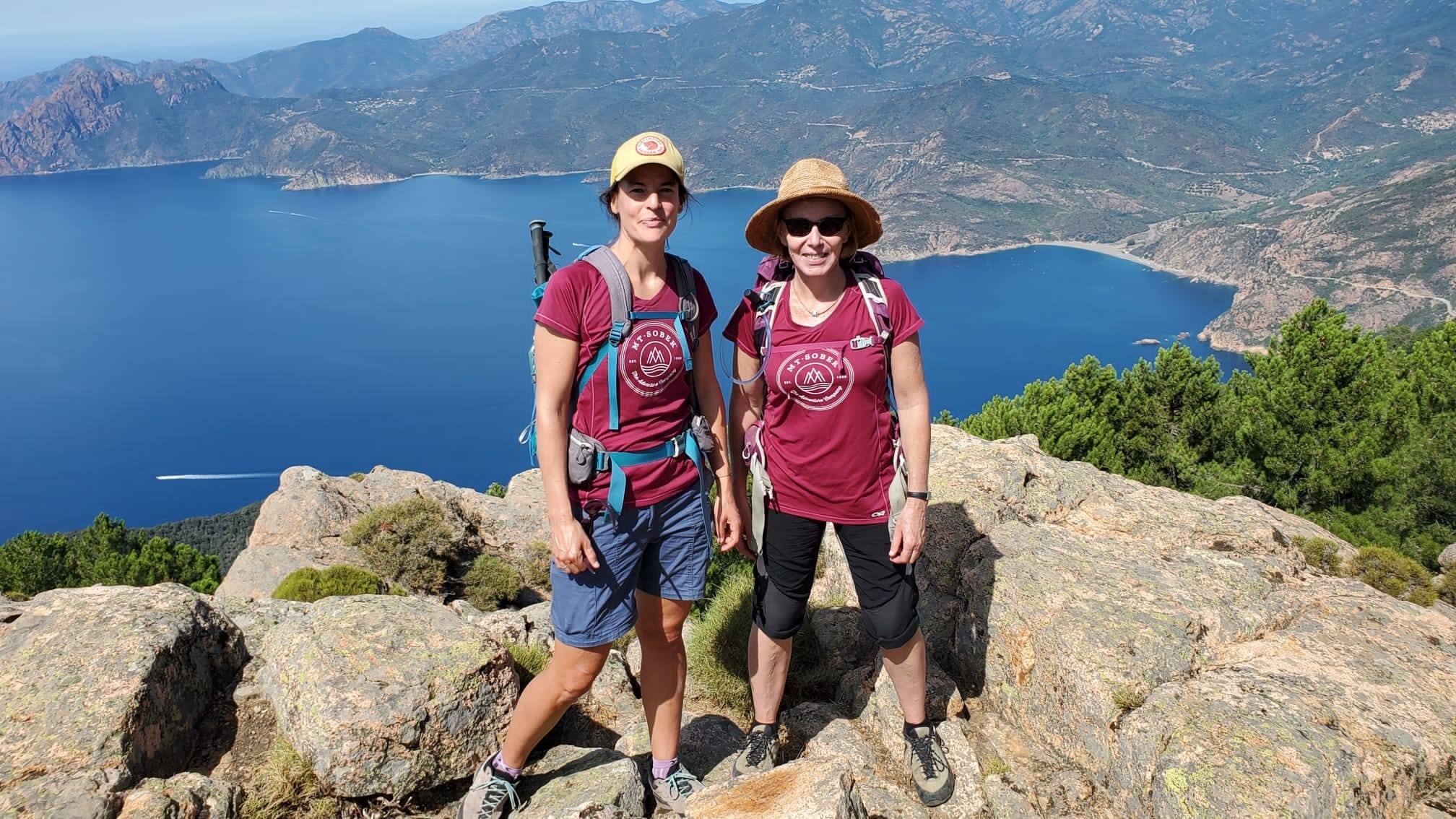 Two women on a thrilling adventure travel, standing on top of a mountain with a breathtaking view of the sea.