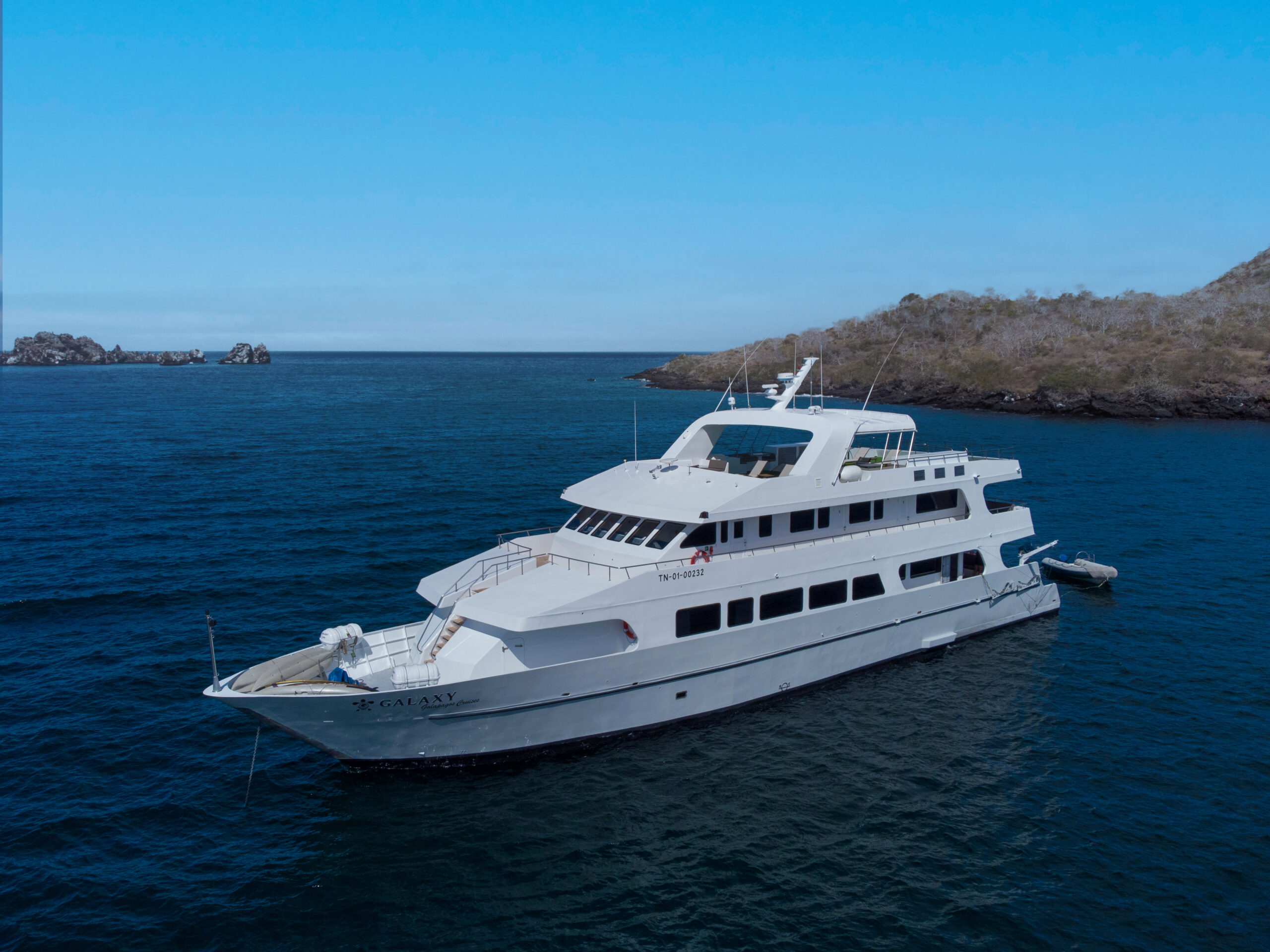A large white yacht is anchored in calm blue waters near a rocky, sparsely vegetated shoreline under a clear sky.