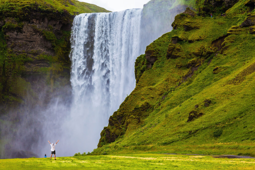 Grand waterfall Skogafoss in Iceland. Tourist in shirt and bandana threw up his hands with delight the beauty of nature. 
