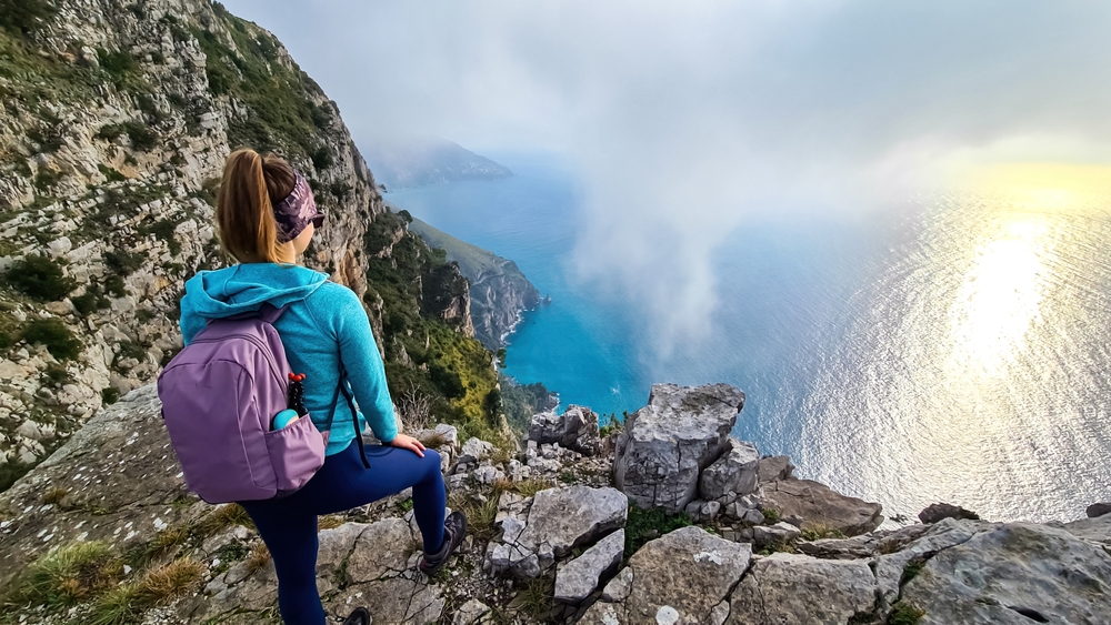 Woman with scenic view from a hiking trail on the coastal driving road of beautiful Amalfi Coast, Campania, Italy, Europe. Riviera coastline at Mediterranean sea. Panoramic curvy road near Positano
