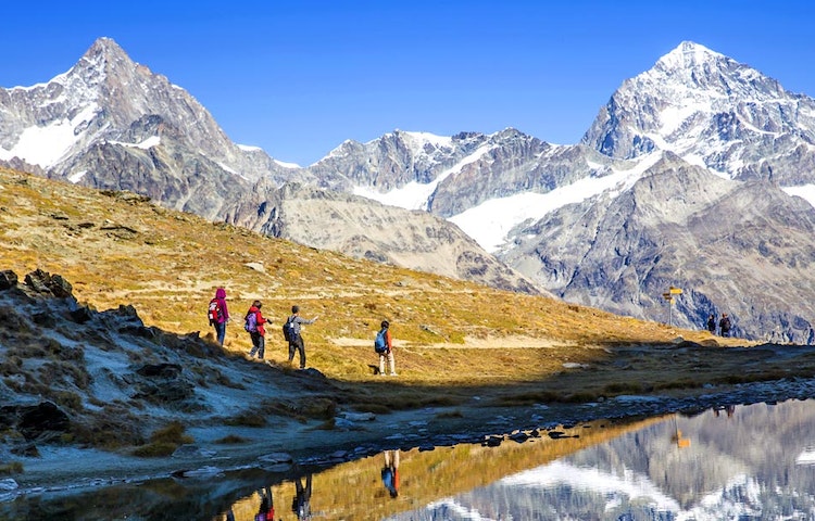 A group of people walking near a lake with mountains in the background.