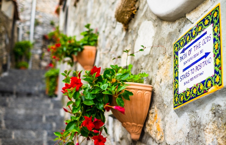A sign on a stone wall with flowers on it, with stairs leading to the Path of Gods in Amalfi Coast, Italy, Europe