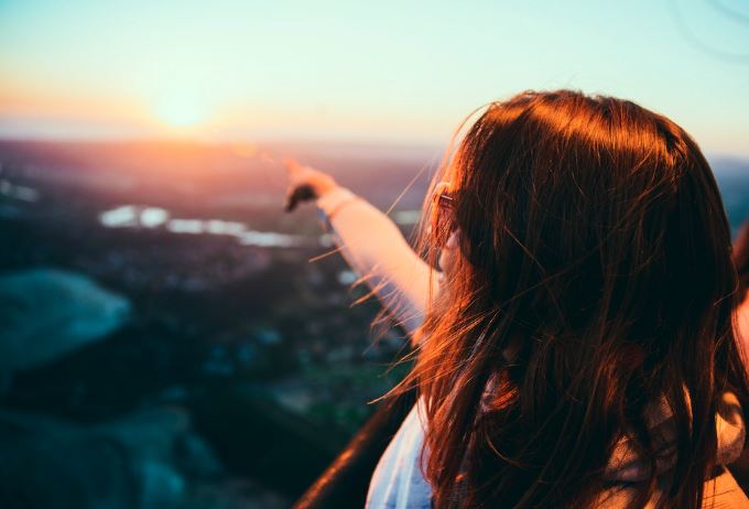 A woman is standing on top of a mountain overlooking a city at sunset.