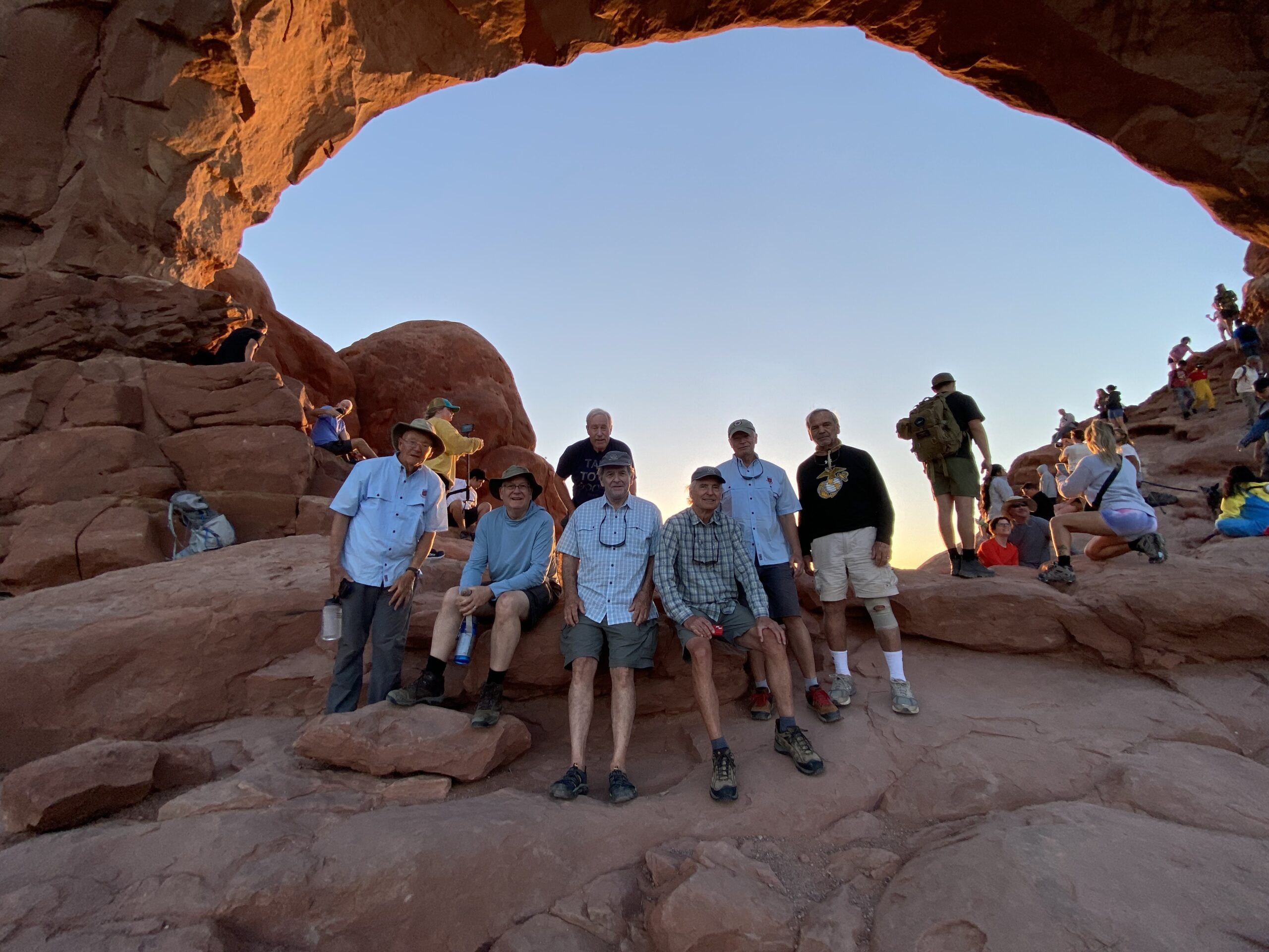 A group of men pose on red rocks beneath a large natural stone arch at sunset, with other visitors in the background.