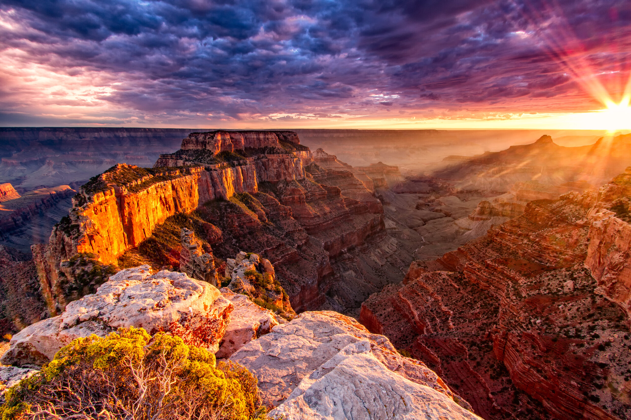 The sun sets over the Grand Canyon, casting warm light on the layered rock formations and dramatic sky filled with clouds.