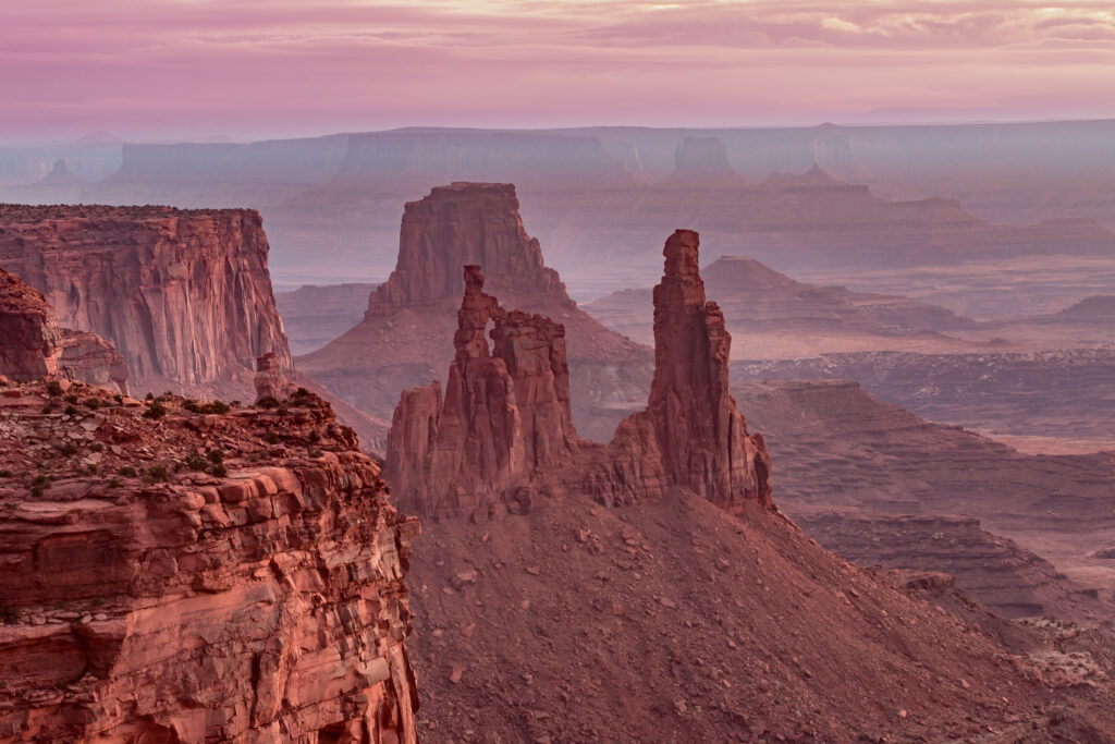 Eroded red rock formations and mesas rise from a desert canyon landscape under a hazy, pinkish sky at sunrise or sunset.