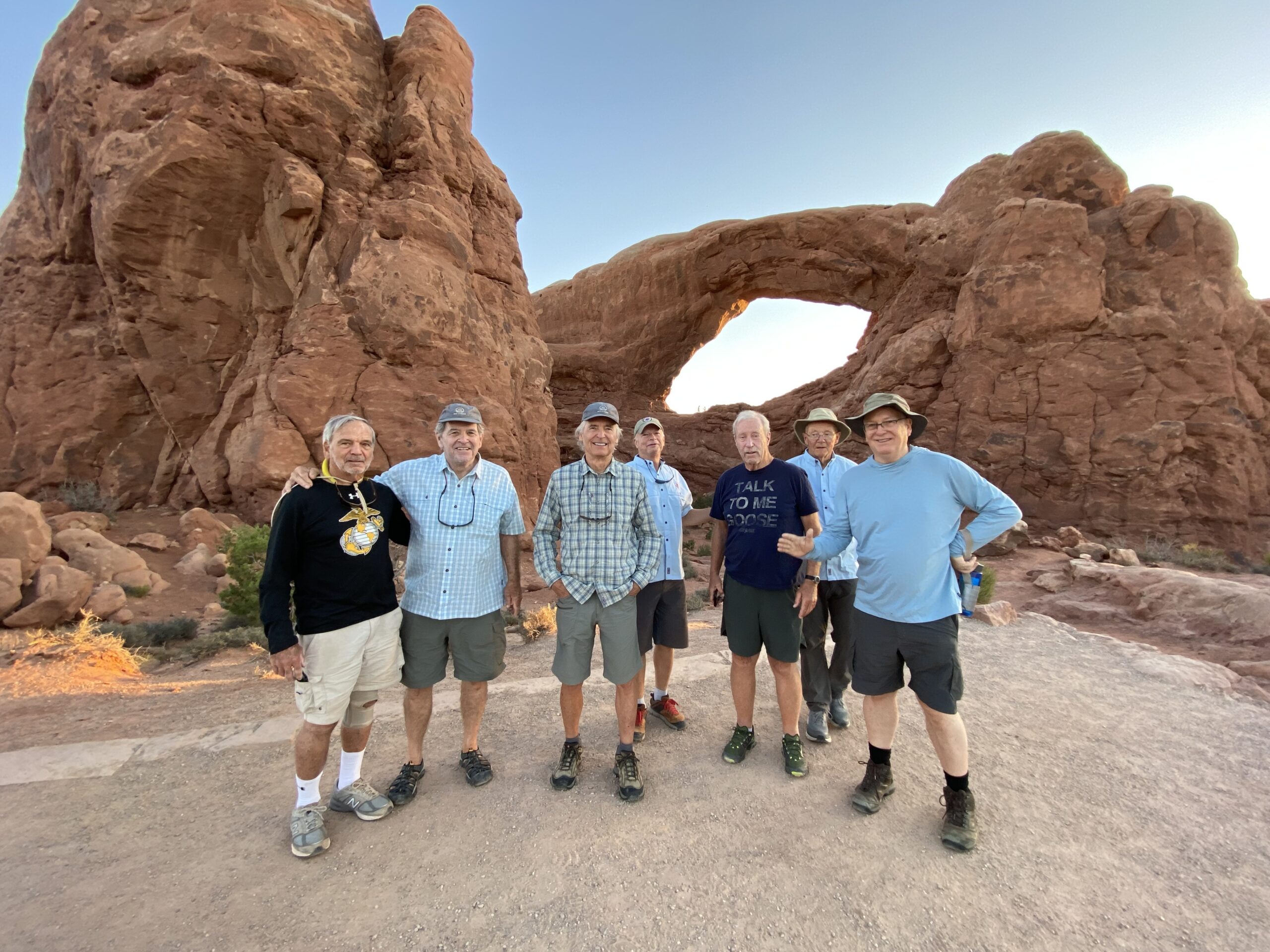 Seven men pose in front of a large rock arch formation in a desert landscape, wearing casual outdoor clothing and hats.