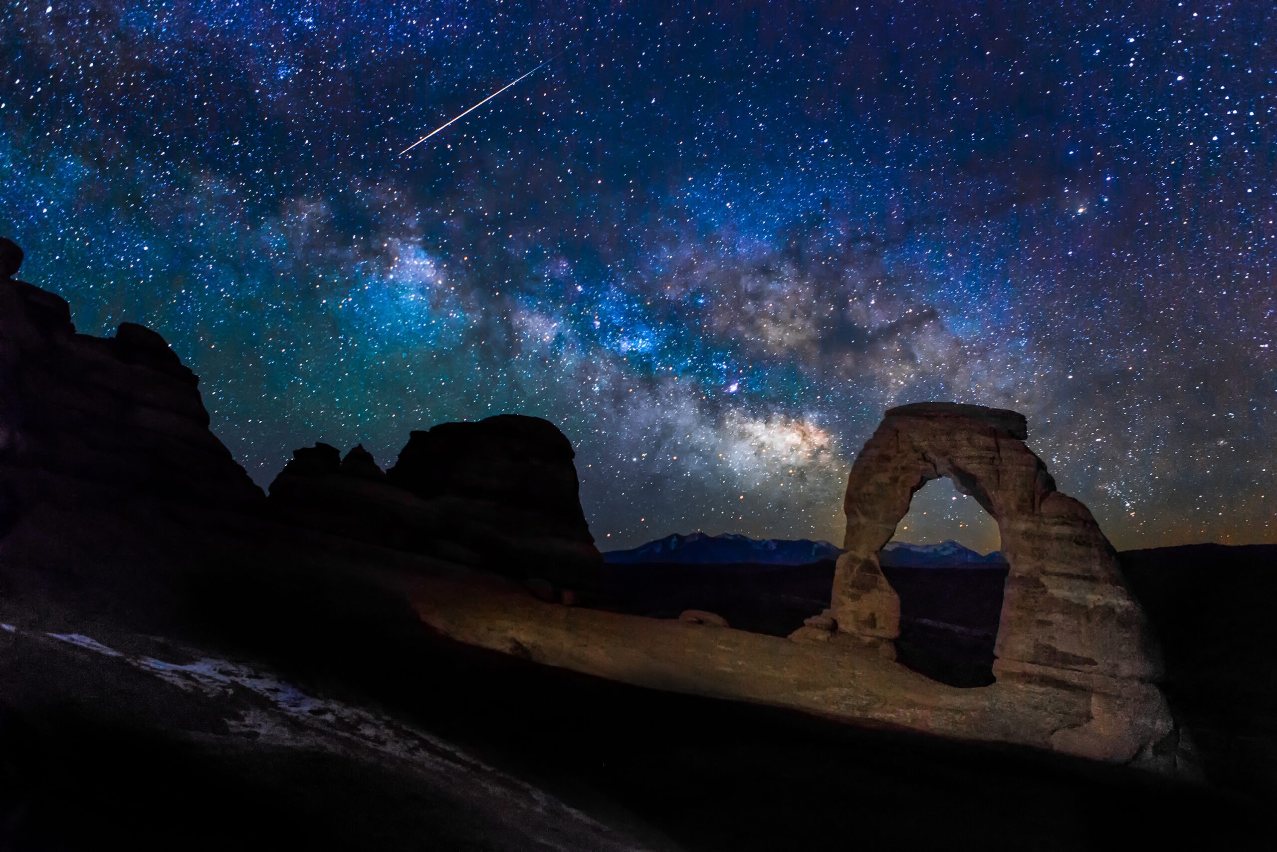 Night sky filled with stars and the Milky Way above Delicate Arch in Arches National Park, with a meteor streaking across the sky.