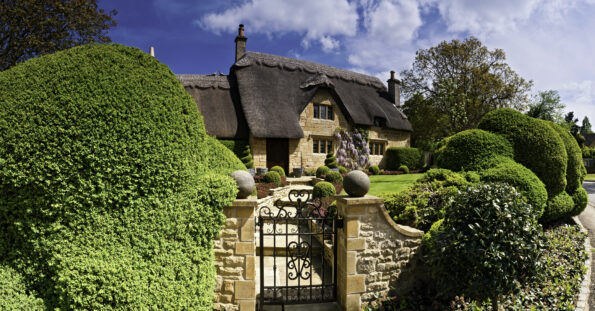 A stone cottage with a thatched roof is surrounded by well-manicured bushes and trees, with a wrought-iron gate in the foreground under a partly cloudy sky.
