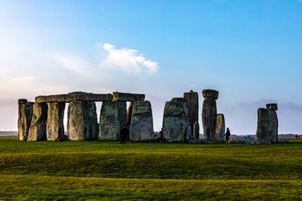 Stonehenge, a prehistoric monument with large standing stones arranged in a circle, sits on a grassy field under a blue sky with a few clouds.