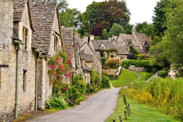 A narrow road curves through a village with old stone cottages, steep roofs, flower-covered walls, and lush greenery on both sides.