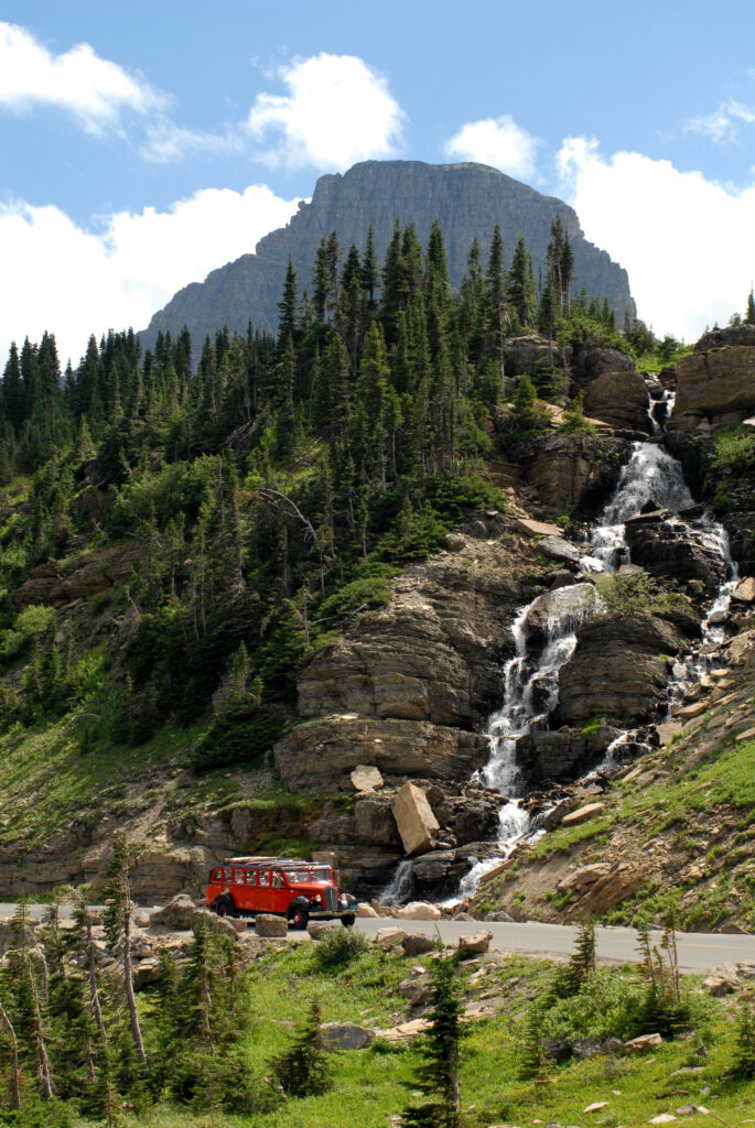 A red tour bus passes by a waterfall at Logan Pass on Glacier Park's Going to the Sun Highway