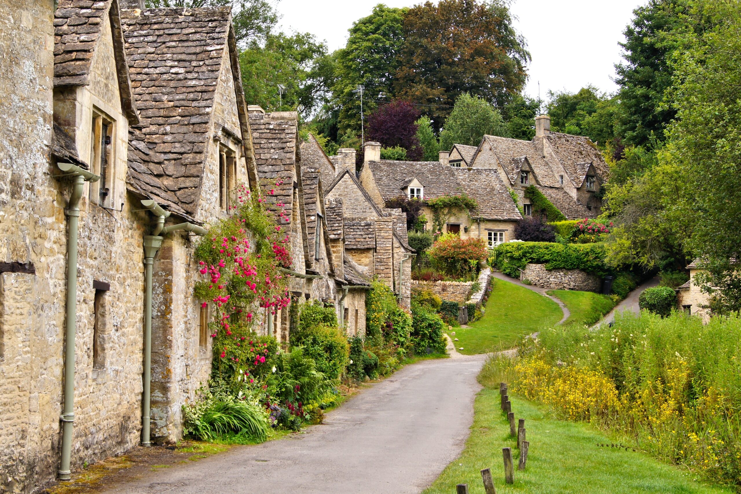 A narrow road curves through a village with old stone cottages, steep roofs, flower-covered walls, and lush greenery on both sides.