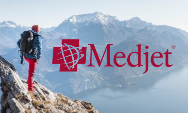 A hiker stands on a rocky mountain ledge overlooking a lake and snowy peaks; the Medjet logo is displayed over the image, highlighting access to essential travel resources.