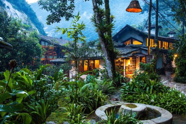A rustic house with glowing windows is surrounded by lush tropical plants and trees, set against a mountainous backdrop at dusk. A stone water feature is in the foreground.