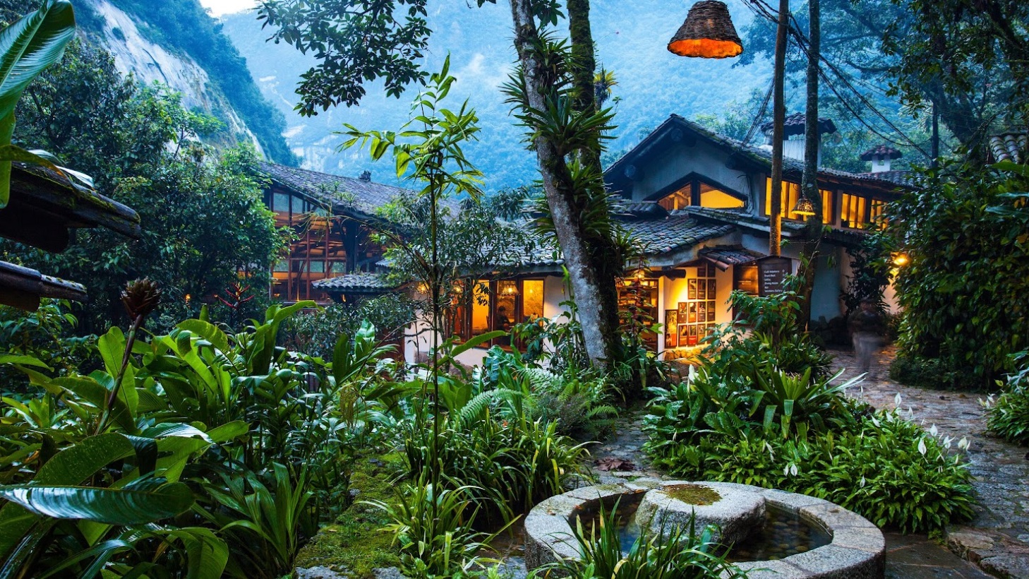 A rustic house with glowing windows is surrounded by lush tropical plants and trees, set against a mountainous backdrop at dusk. A stone water feature is in the foreground.