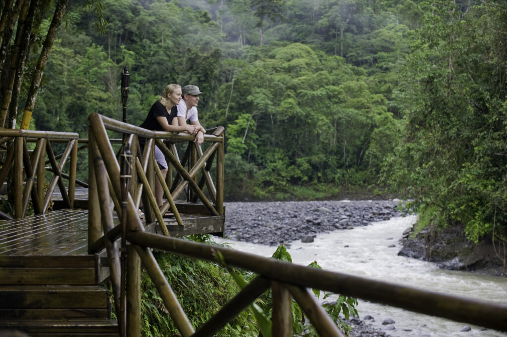 two female and male travelers standing in front of Rios Lodge looking at the Pacuare River and hearing sounds of toucans