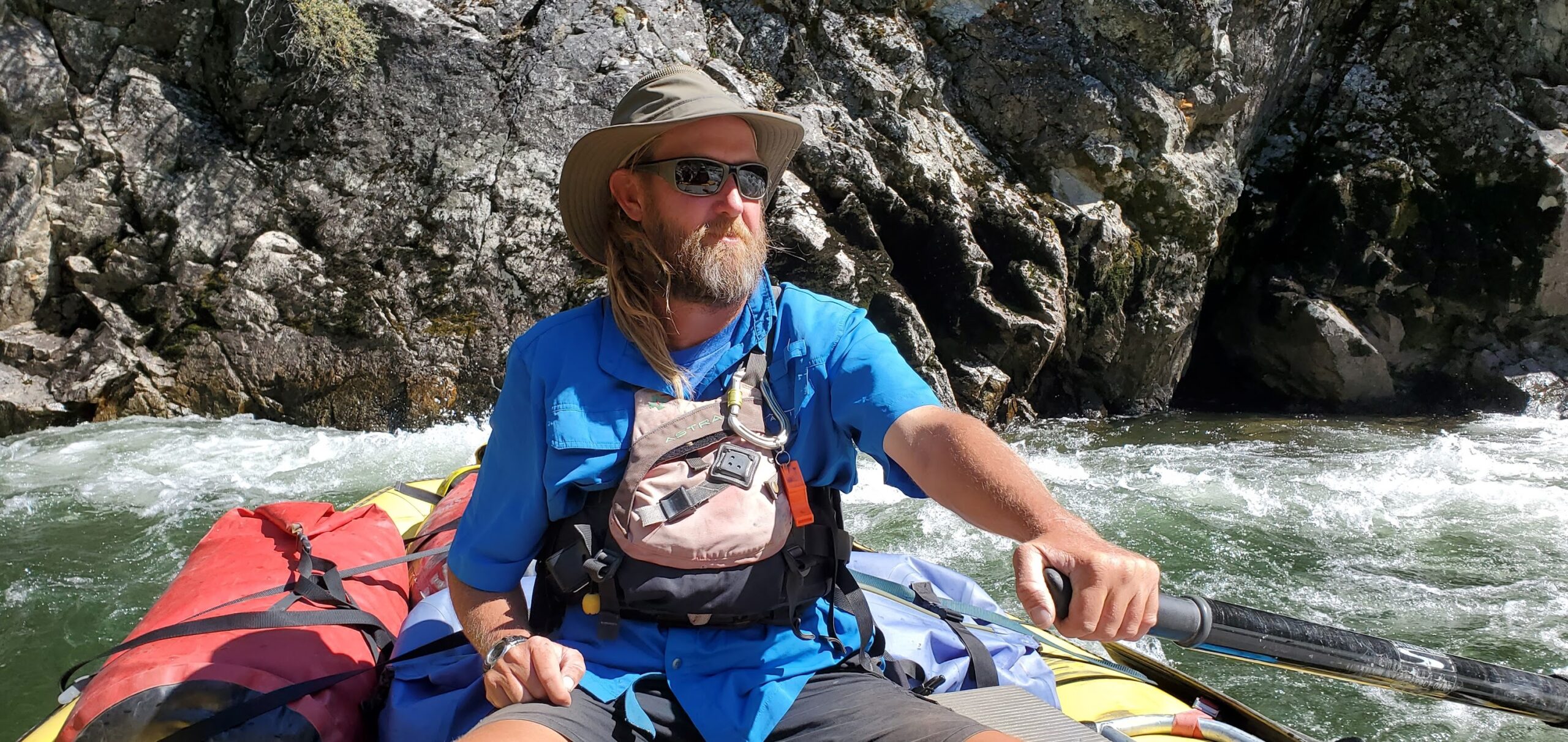 A person wearing a hat, sunglasses, and a life vest paddles a raft through rocky cliffs, capturing the thrill of Idaho River Rafting.