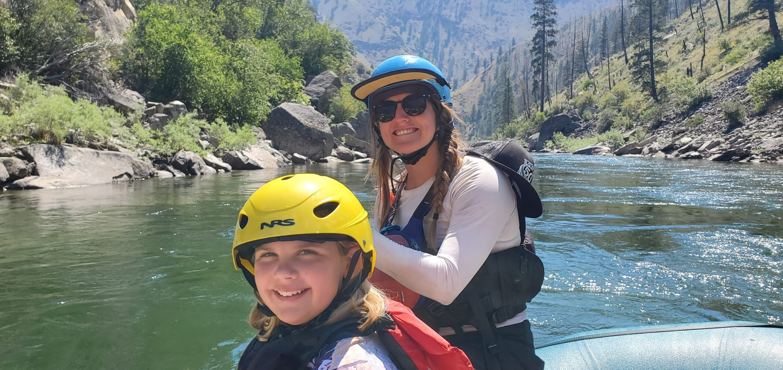 Two people wearing helmets and life jackets sit on a raft, smiling, as they enjoy an unforgettable Idaho River Rafting adventure with scenic rivers and forested mountains in the background.