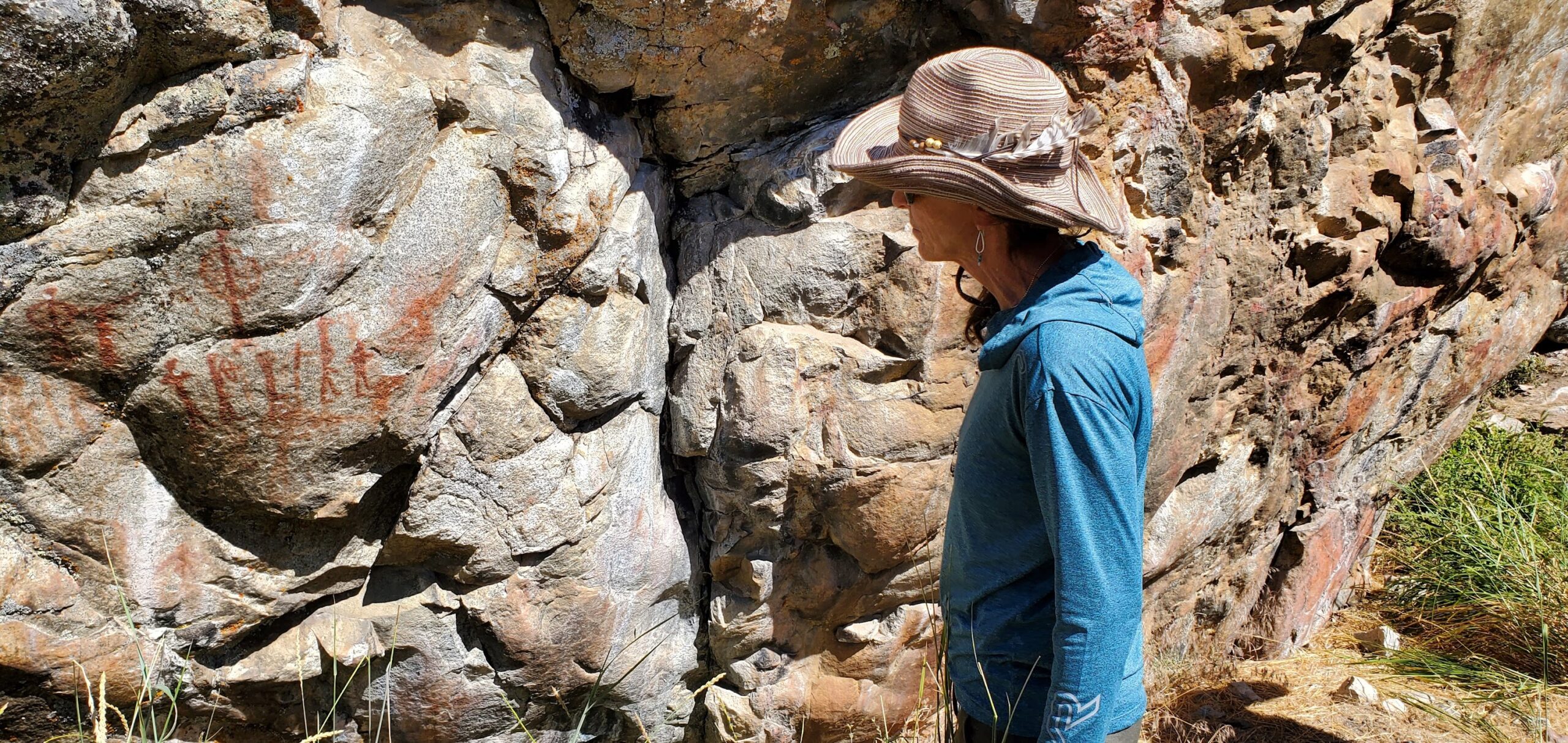 Person wearing a hat and blue shirt stands next to a large rock face with red ochre markings, outdoors in a grassy area—capturing the adventurous spirit of Idaho River Rafting.