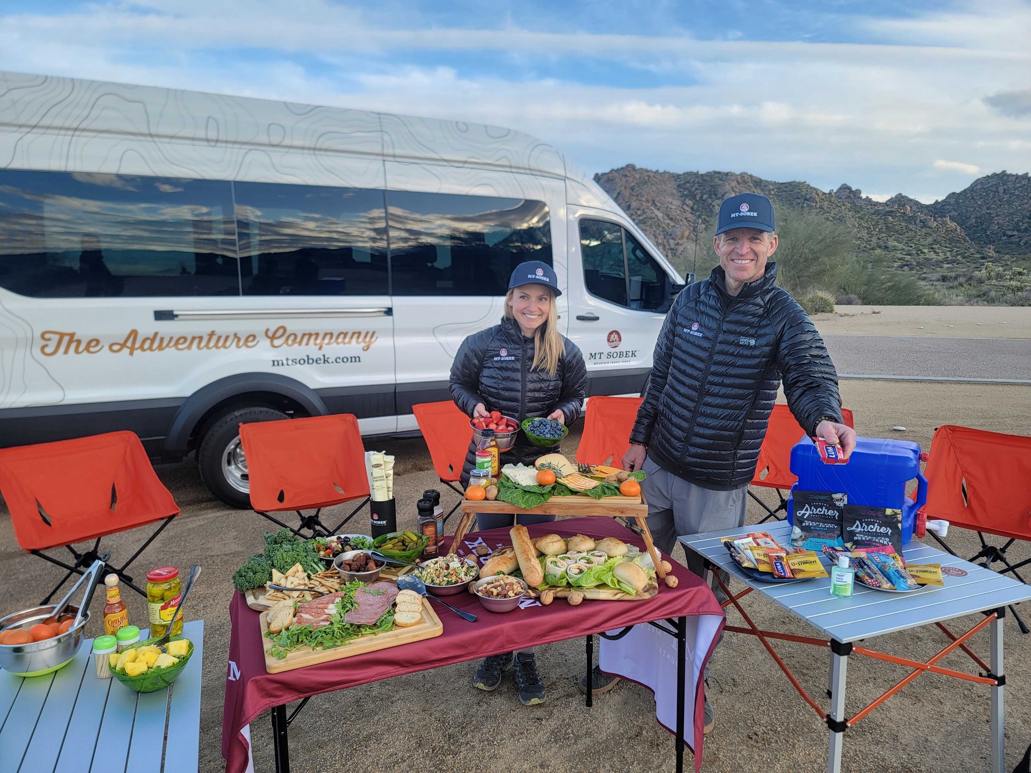 Two people stand beside tables with assorted food and drinks, set up outdoors near a van and red chairs—an ideal scene for corporate group retreats, with mountains and desert landscape in the background.