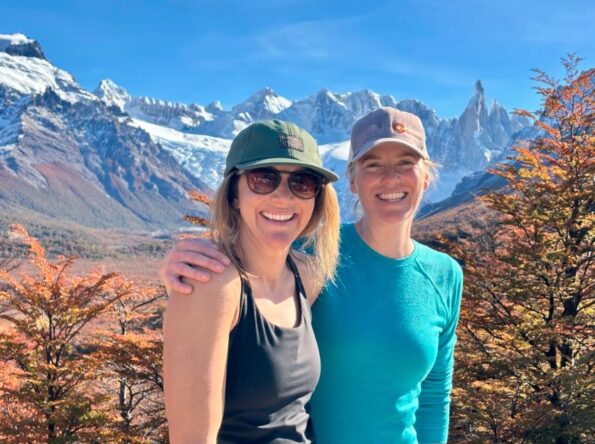 Two women in casual outdoor clothing smile in front of snow-capped mountains and autumn-colored trees under a clear blue sky.