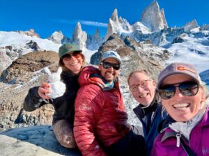 Four people in outdoor gear smile for a selfie on a rocky slope with snowy mountains in the background; one holds a small snow sculpture.