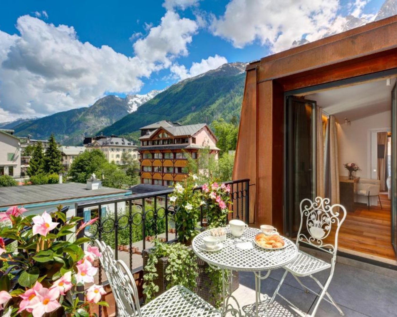 A balcony with a table and two chairs set for breakfast overlooks charming buildings and a mountainous landscape at the Hotel des Alpes with a blue sky and clouds.