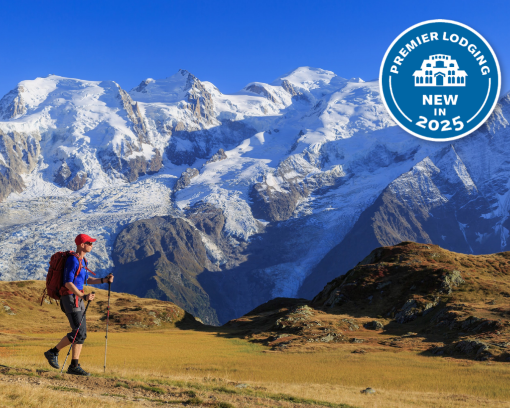 A male hiker walks on a trail beneath snow-capped mountains in the Alps in Europe. A badge on the top-right corner reads "Premier Lodging, New in 2025.