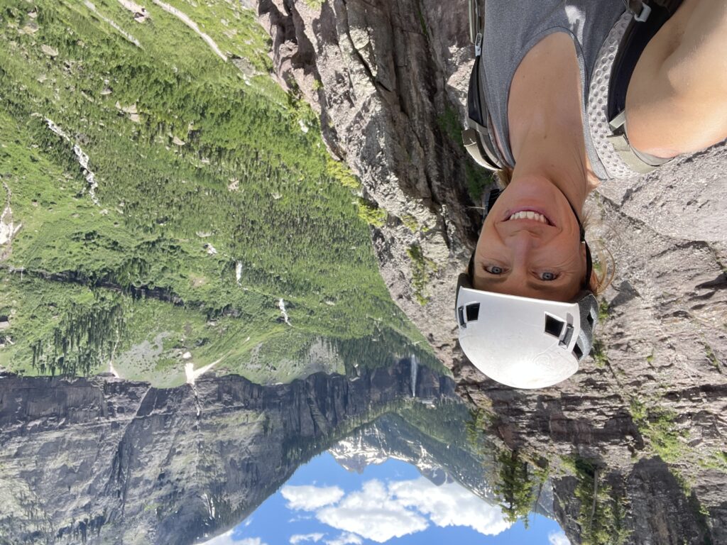 A person wearing a helmet and backpack smiles while standing on a rocky ledge with a scenic valley and distant mountains in the background in Denver, Colorado