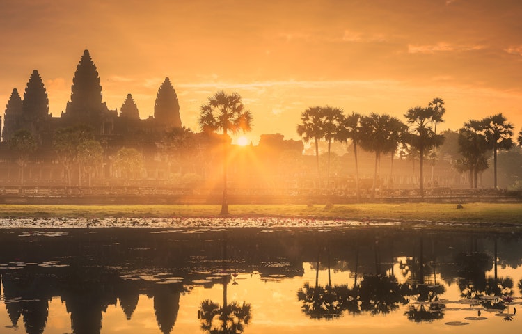 A sunrise illuminates Angkor Wat temple in Cambodia, casting a golden glow over the surrounding palm trees and reflecting in a calm lake.