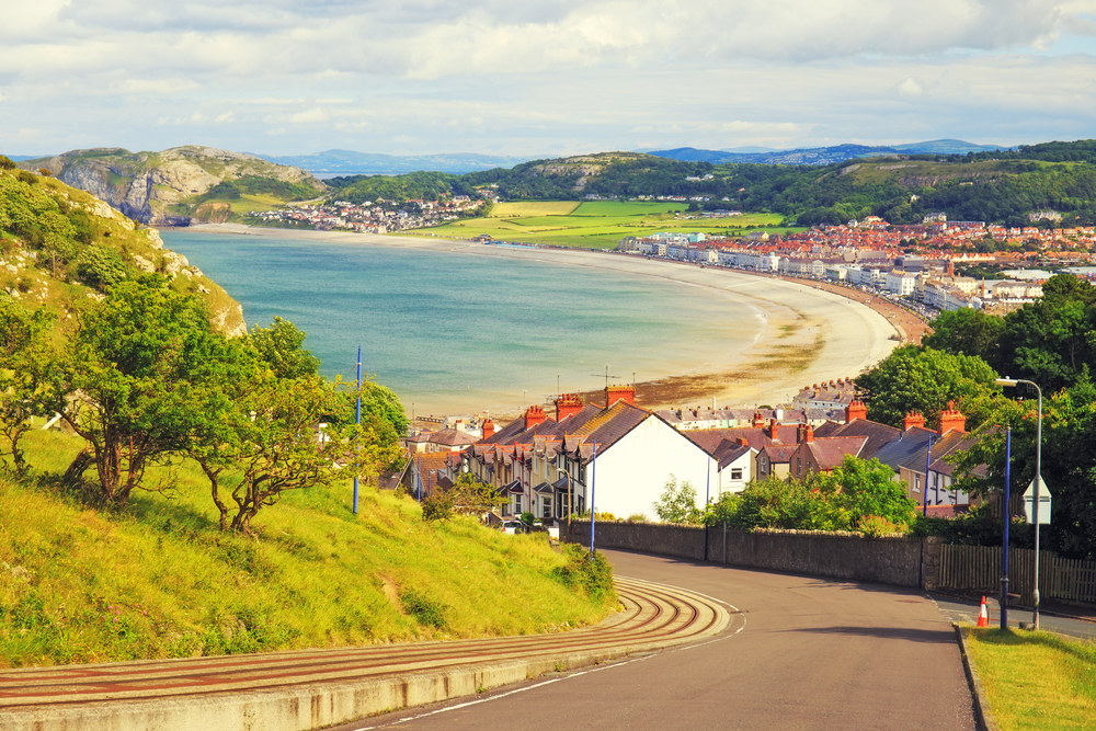 A scenic coastal town features curved roads, residential houses, and a long sandy beach with hills and greenery in the background on a partly cloudy day in England, Lake District.