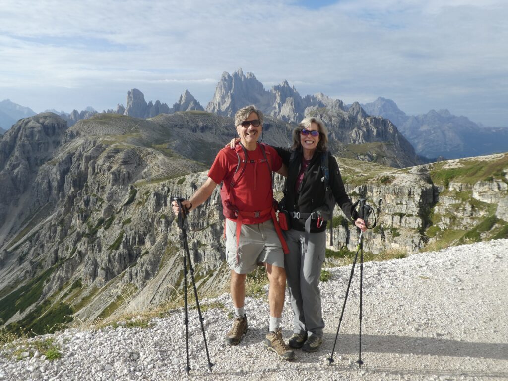 Two active hikers a couple with trekking poles stand on a rocky path in front of steep Dolomites mountain peaks under a partly cloudy sky in Italy, Europe