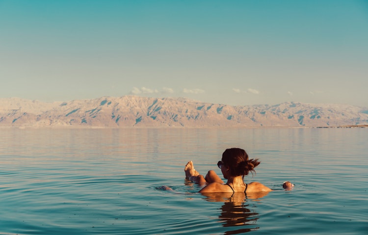 A person floating on their back in a calm body of water in the Dead Sea with a distant mountain range in the background under a clear blue sky in Jordan in the Middle East