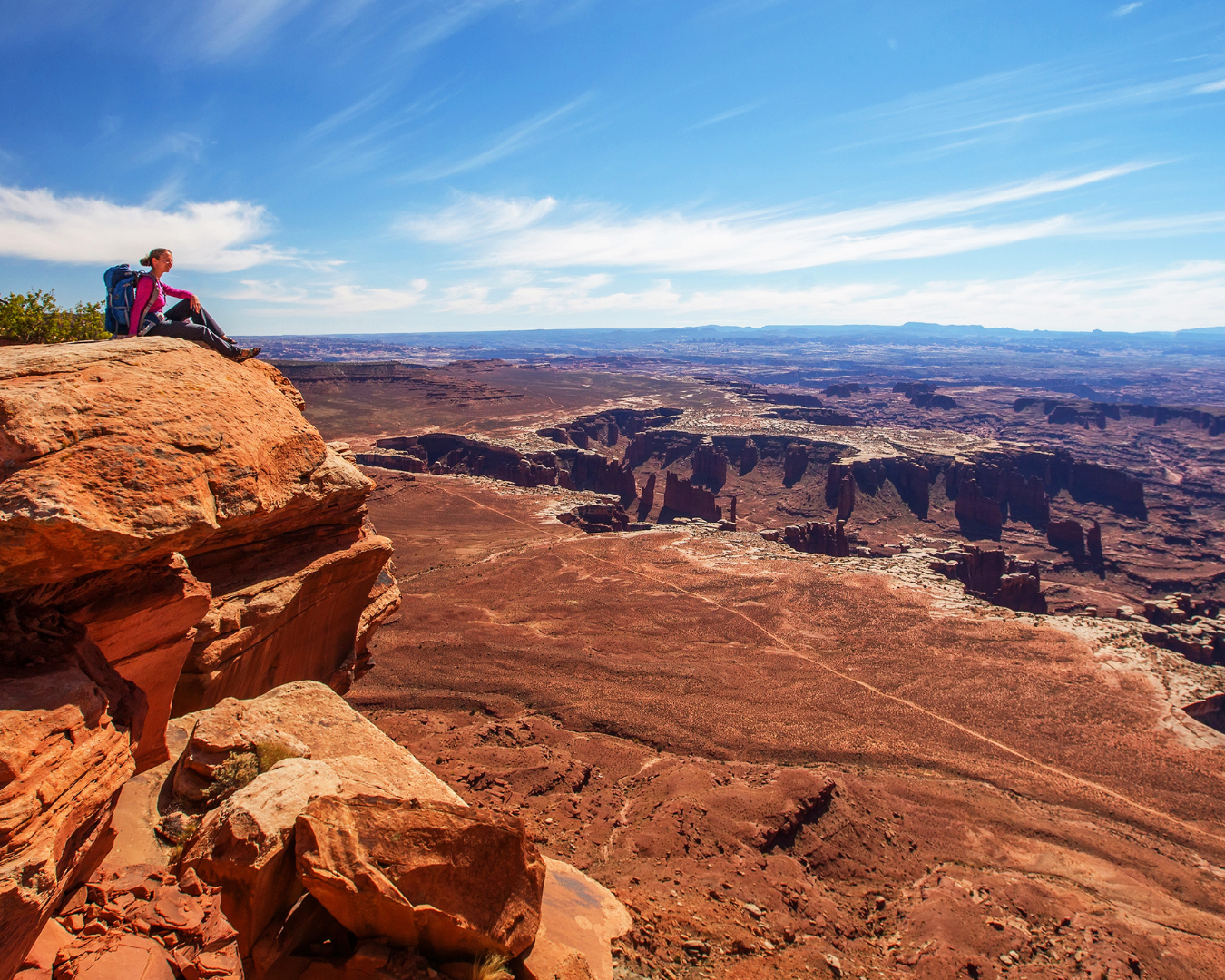 Female hiker sitting on the edge of a rock cliff overlooking a vast desert canyon landscape of Utah's arches and hoodoos under a clear sky at Canyonlands National Park