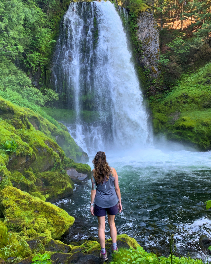 A person in outdoor attire stands on a mossy rock facing a large waterfall surrounded by lush greenery in a forest setting