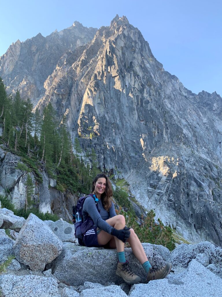 A person with long hair and a backpack is sitting on rocks with a mountainous backdrop in the pacific northwest. They are wearing hiking gear and are smiling at the camera.