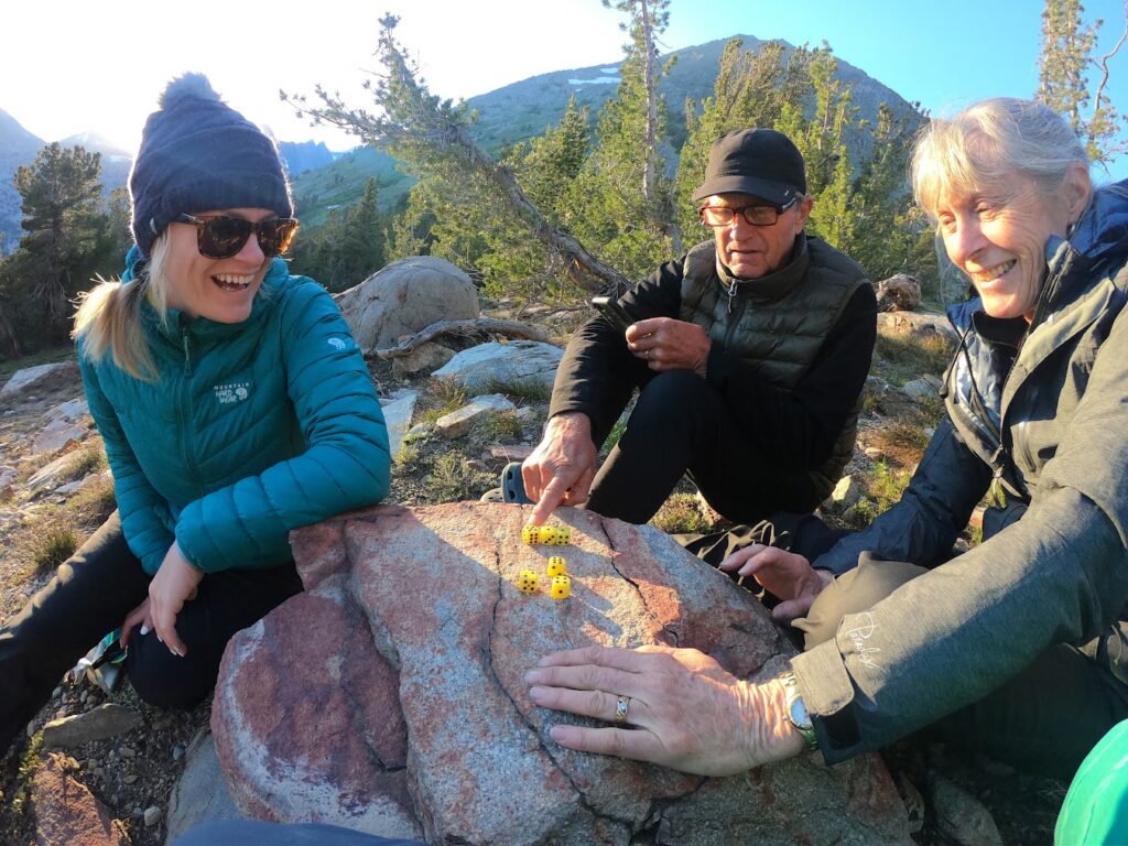 Three people sitting outdoors on rocky terrain, playing a game with yellow dice on a large rock. Mountain and trees visible in the background.