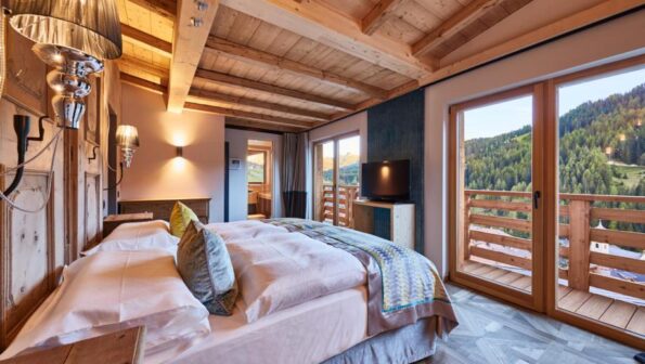 A cozy premier lodging option featuring a wooden ceiling, a double bed adorned with pastel linens, wall-mounted lamps, a flat-screen TV, and large glass doors that open to a breathtaking mountain view.