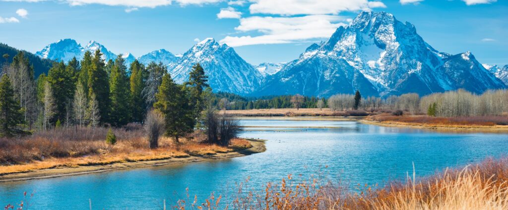 A calm river flows through a forested area with tall, snow-capped mountains in the background under a partly cloudy sky—an ideal scene for those seeking the best US adventure tours.