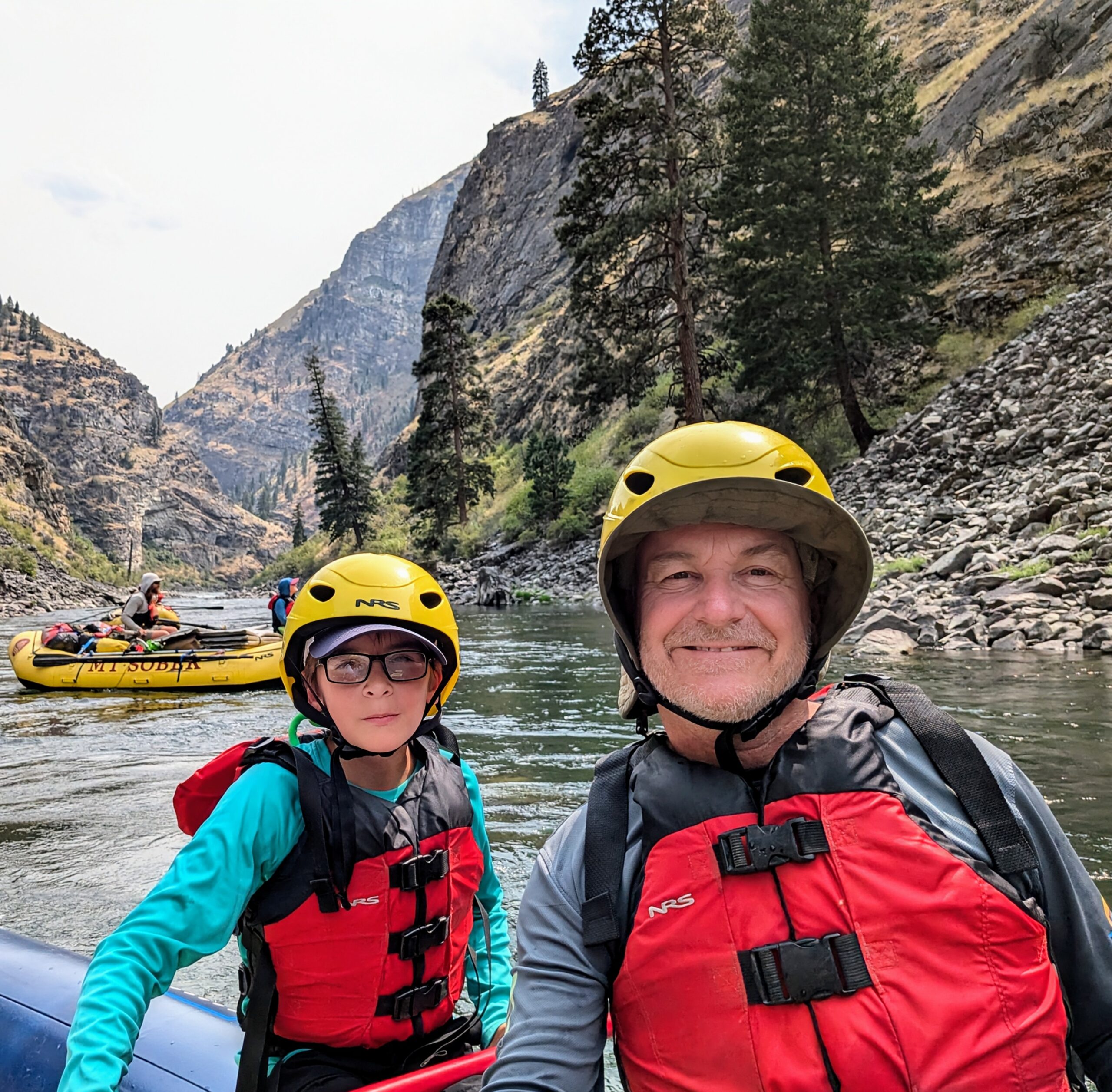 Two people in helmets and life jackets sit in a raft on a river, surrounded by mountains and trees; another raft is visible in the background.