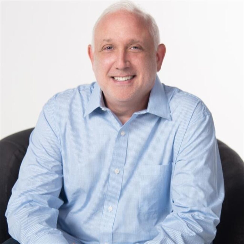 A man with short white hair, wearing a light blue button-up shirt, sits on a dark chair and smiles at the camera against a plain white background.