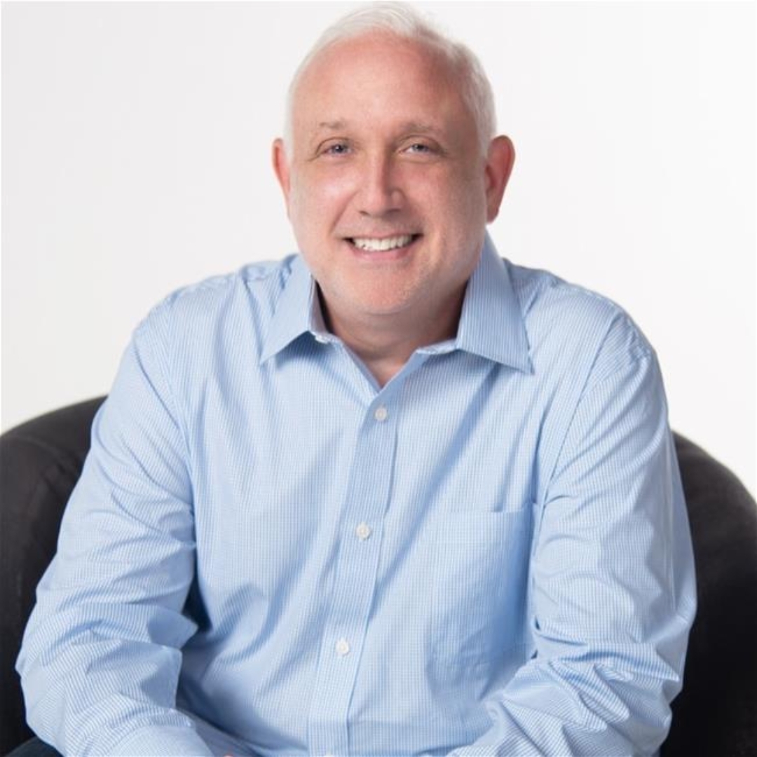 A man with short white hair, wearing a light blue button-up shirt, sits on a dark chair and smiles at the camera against a plain white background.