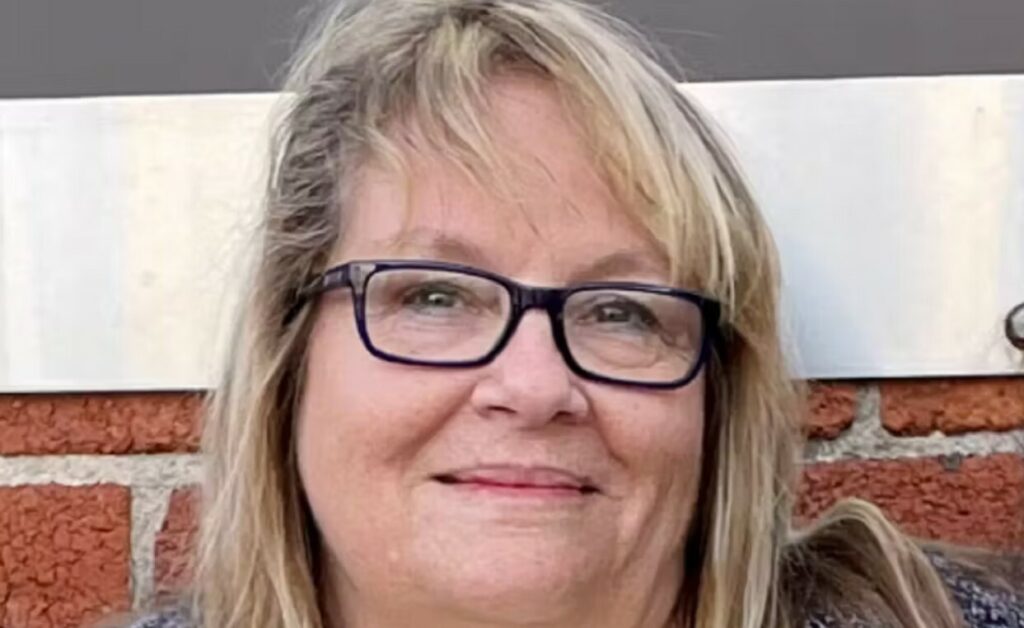 A woman with blonde hair and glasses smiles slightly while standing in front of a brick wall and a gray metal panel.
