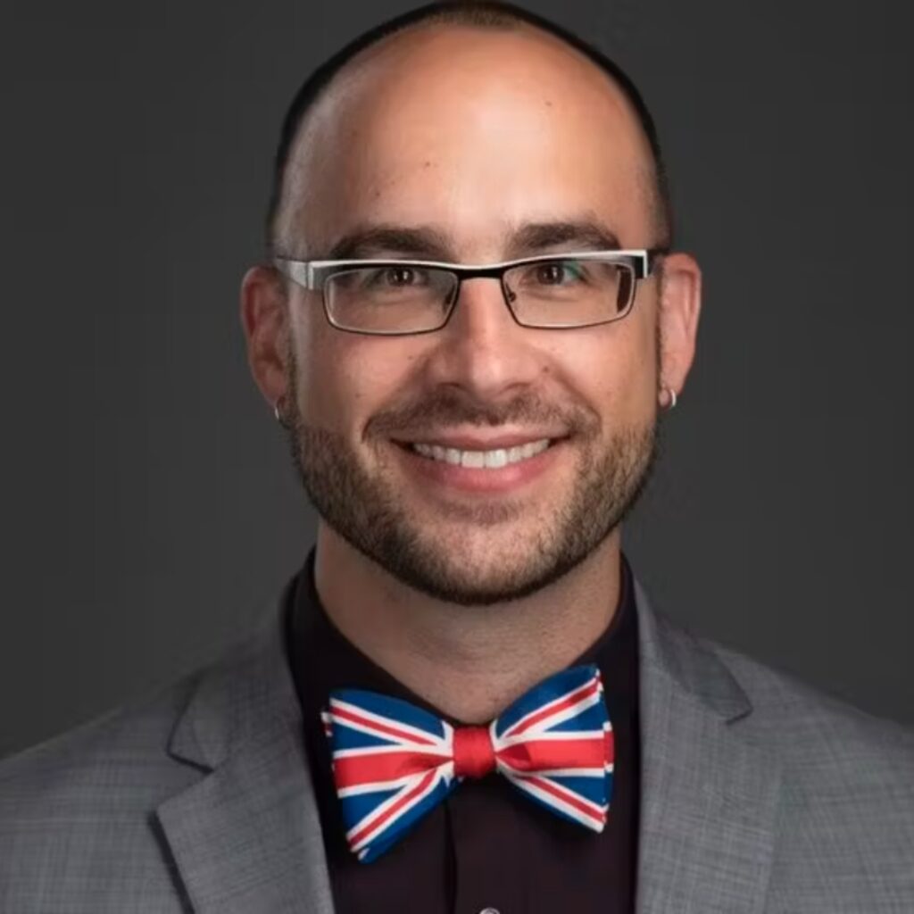 A man with glasses and a beard, wearing a gray suit, dark shirt, and a Union Jack bow tie, smiles against a dark background.