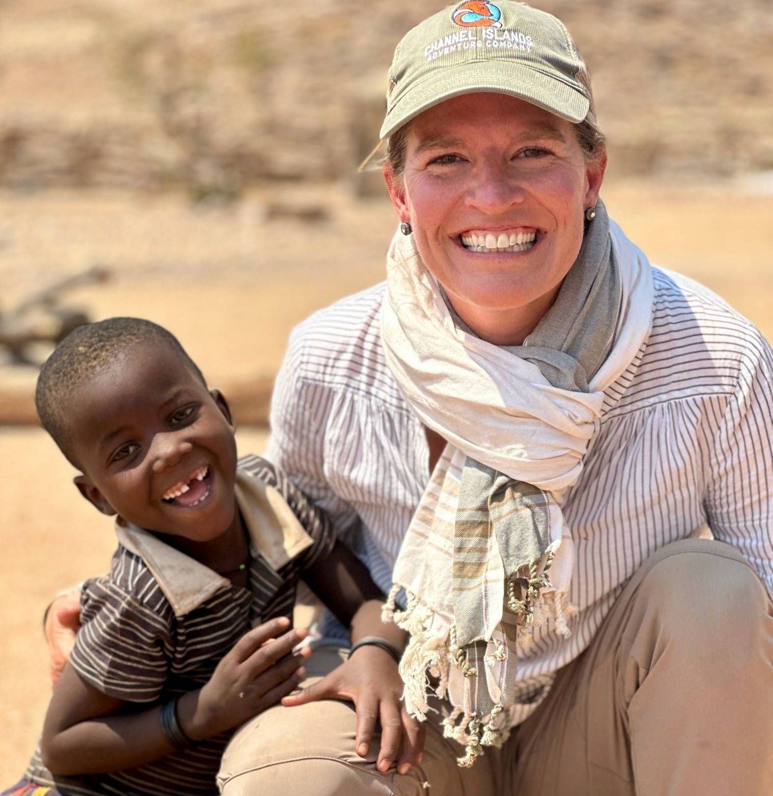 A smiling woman in a cap and scarf crouches next to a young boy in a striped shirt, both smiling at the camera in an outdoor setting.