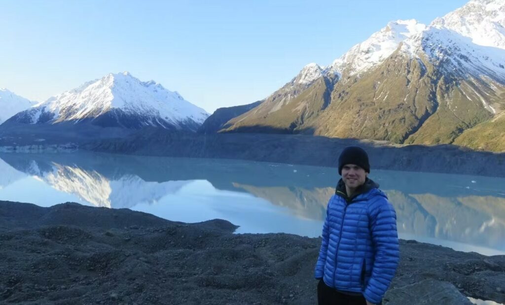 A person wearing a blue jacket and black beanie stands by a calm lake with snow-capped mountains reflected in the water under a clear sky.