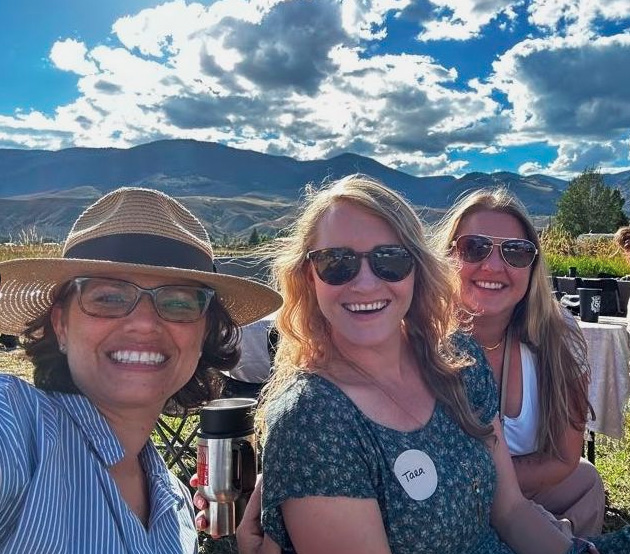 Three women sit outdoors smiling, wearing sunglasses; one wears a striped shirt and hat, another has a name tag reading "Tara." Mountains and clouds are visible in the background.