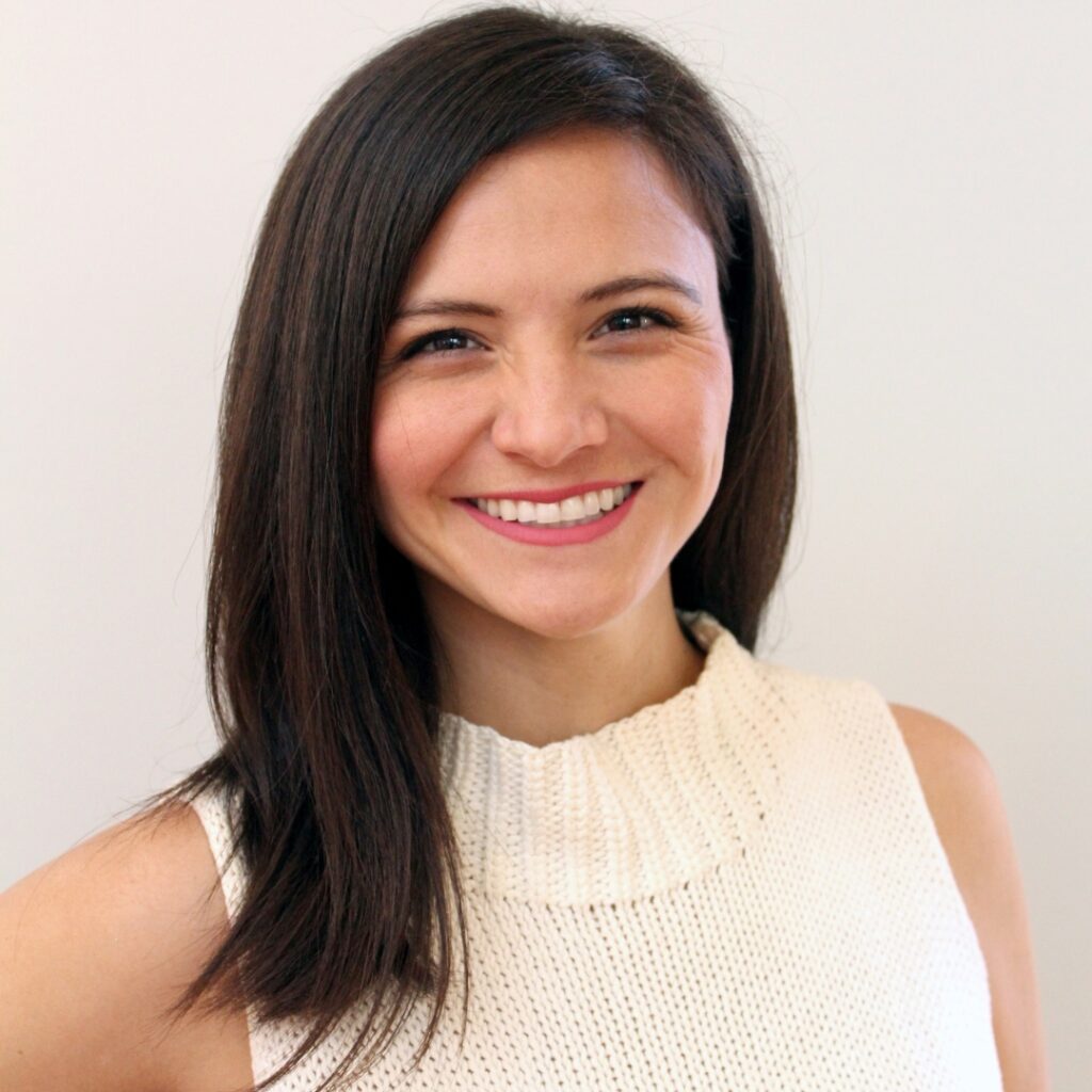 A woman with straight, dark hair smiles at the camera. She is wearing a sleeveless, cream-colored knit top and standing against a plain, light background.
